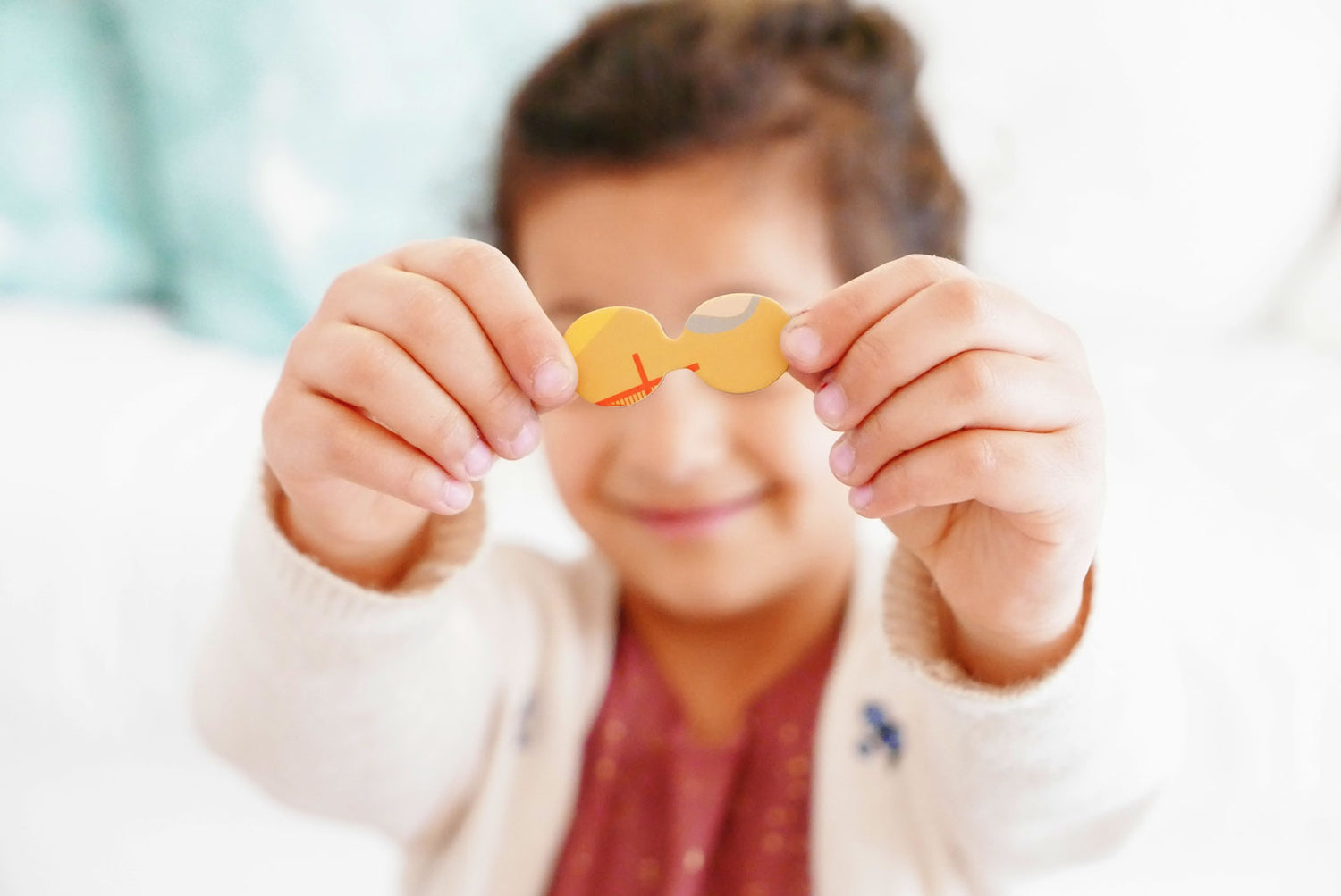 Portrait photograph of a young girl smiling while holding up a custom puzzle piece shaped like a pair of eyeglasses