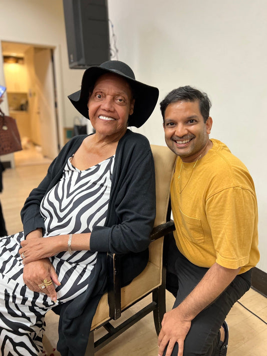 Miss Major Griffin-Gracy, seated, wears a black hat and black and white dress, to her right is Rajiv kneeling wearing a yellow shirt