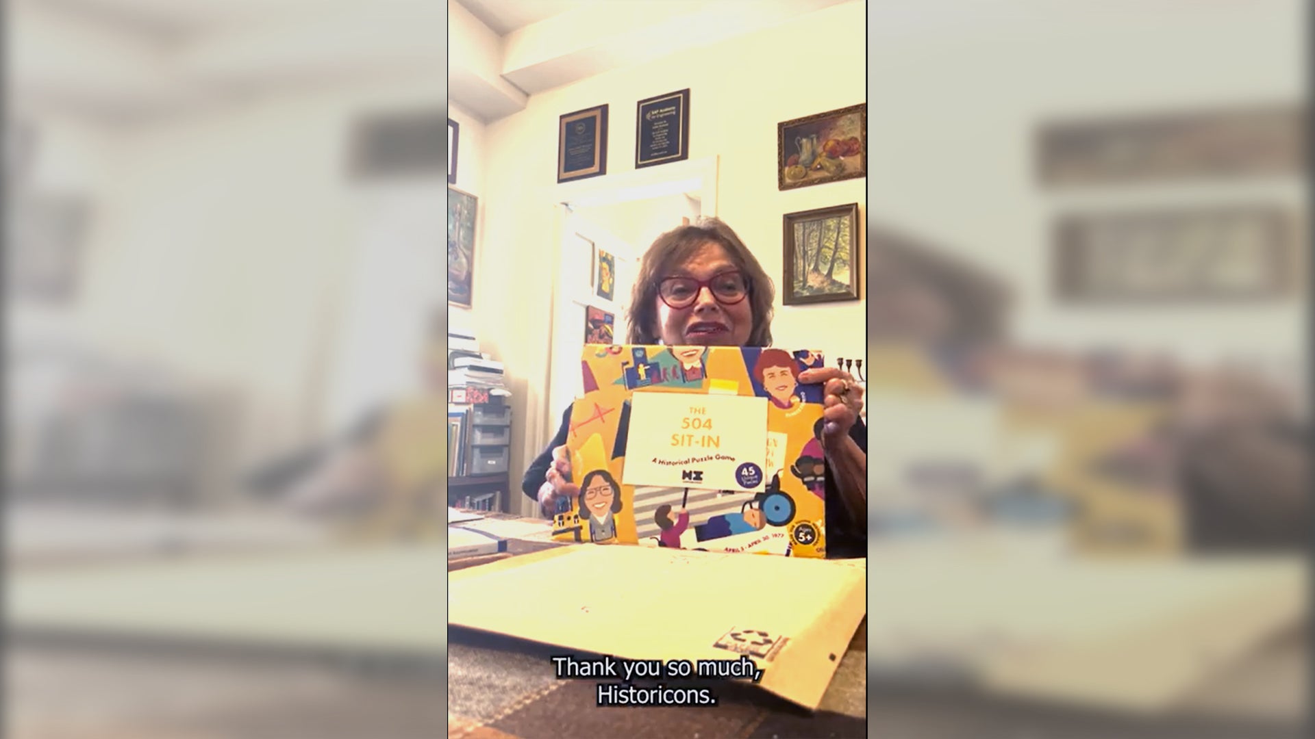 Load video: Disability rights icon Judy Heumann sits at a table and unboxes The 504 Sit-In puzzle game from Historicons. She removes the puzzle from a sleeve and talks about the people featured on it, comments on the colorful yellow puzzle, and thanks Historicons for bringing disability history to the forefront.